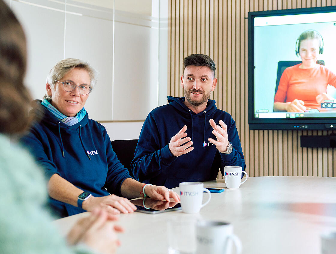 Besprechungssituation mehrerer Personen, ein Mann erklärt etwas. Im Hintergrund ist eine Person per Video zugeschaltet.