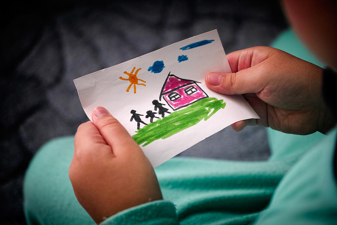 Kind das ein selbstgemaltes Bild in den Händen hält, auf dem eine Familie vor einem Haus zu sehen ist.
