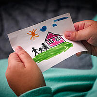 Kind das ein selbstgemaltes Bild in den Händen hält, auf dem eine Familie vor einem Haus zu sehen ist.