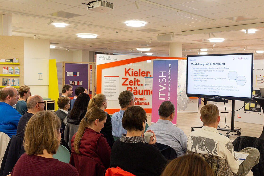 Teilnehmende des Netzwerktreffens Digitale Langzeitarchivierung sitze auf Stühlen vor einer Präsentation in einem Konferenzraum.